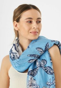 Écharpe bleu clair arborant des motifs floraux et de papillons en bleu marine. Fabriquée dans un tissu doux et texturé. Portée lâchement autour du cou.