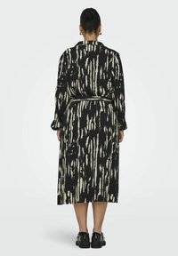 Robe longue noire avec un motif abstrait crème, manches longues, taille à nouer et coupe ample. Portée avec des chaussures noires à semelles épaisses.
