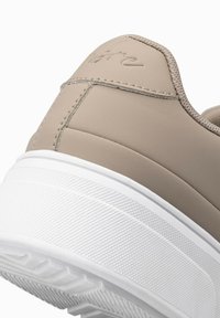 Baskets synthétiques beige avec une semelle en caoutchouc blanc texturé. Présente des accents de couture et un panneau de talon lisse embossé du nom de la marque.