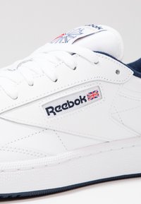 Baskets blanches Reebok avec des accents bleu marine, des lacets, un patch logo et un drapeau Union Jack sur le côté et la languette.