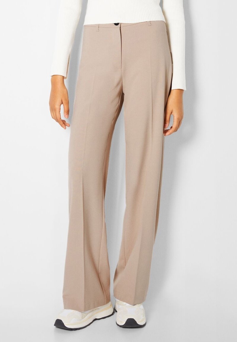 Femme portant un pantalon beige à jambes larges, un haut blanc à manches longues et des baskets blanches, debout devant un fond clair uni.