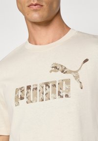 Detailní záběr osoby v světle béžovém tričku s béžovým a hnědým maskáčovým vzorem loga Puma a textem na hrudi.