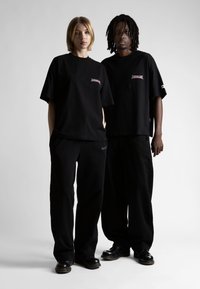 Dvaja jednotlivci majú na sebe zladené čierne nadmerné tričká s grafickým dizajnom a nohavice. Tkanina vyzerá jemne, má minimálne akcenty a uvoľnený strih.