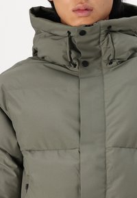 Olive green puffer jacket featuring a high collar, toggle drawstrings, and snap closures. Smooth, shiny exterior with a slightly quilted design.