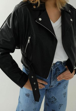 Czarna skórzana kurtka typu cropped z srebrnymi suwakami, na białej koszulce z dekoltem w serek oraz niebieskich dżinsach z nadrukami i sloganami w stylu ręcznego pisma.