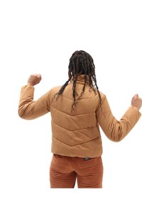 Chaqueta de plumas marrón con cuello alto y patrón de costura tipo chevron. El modelo lleva pantalones de pana, visto desde atrás con los brazos levantados.