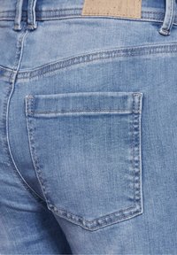 Light blue denim jeans with a back pocket and contrasting stitching. Texture appears soft with slight fading and a rectangular leather label.