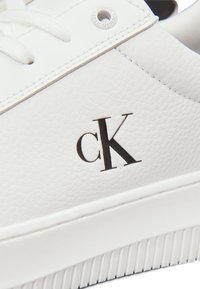 Sneaker in pelle bianca con superficie testurizzata, punta tonda e logo "cK" nero sul lato. Include lacci piatti bianchi e suola testurizzata.