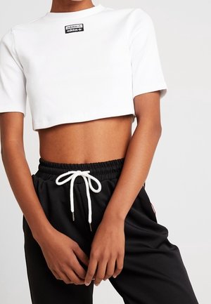 Personne portant un t-shirt court blanc Adidas et un pantalon noir à cordon avec les mains posées près de la taille.