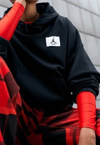 Felpa nera con tasca a marsupio, dotata di un'etichetta con logo bianco. Il modello indossa maniche lunghe rosse e pantaloni rossi a fantasia, creando un contrasto visivo.