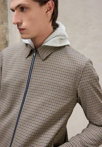 Kollane ja tumesinine houndstooth-mustriga jakk, millel on lukuga kinnis ja hall kapuutsivooder. Omab struktureeritud kraed ja tekstureeritud viimistlust.