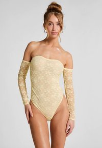 Kanten body in lichtgeel met lange doorschijnende mouwen, bloemenpatroon en bandloze ontwerp; gladde textuur en aansluitend silhouet.