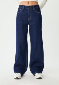 Tamno plave široke traperice od denima; imaju gustu šavovu obradu, dugme za zatvaranje i dizajn s pet džepova. Uparen s bijelim tenisicama.