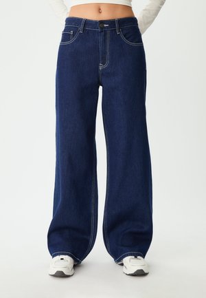 Tamno plave široke traperice od denima; imaju gustu šavovu obradu, dugme za zatvaranje i dizajn s pet džepova. Uparen s bijelim tenisicama.