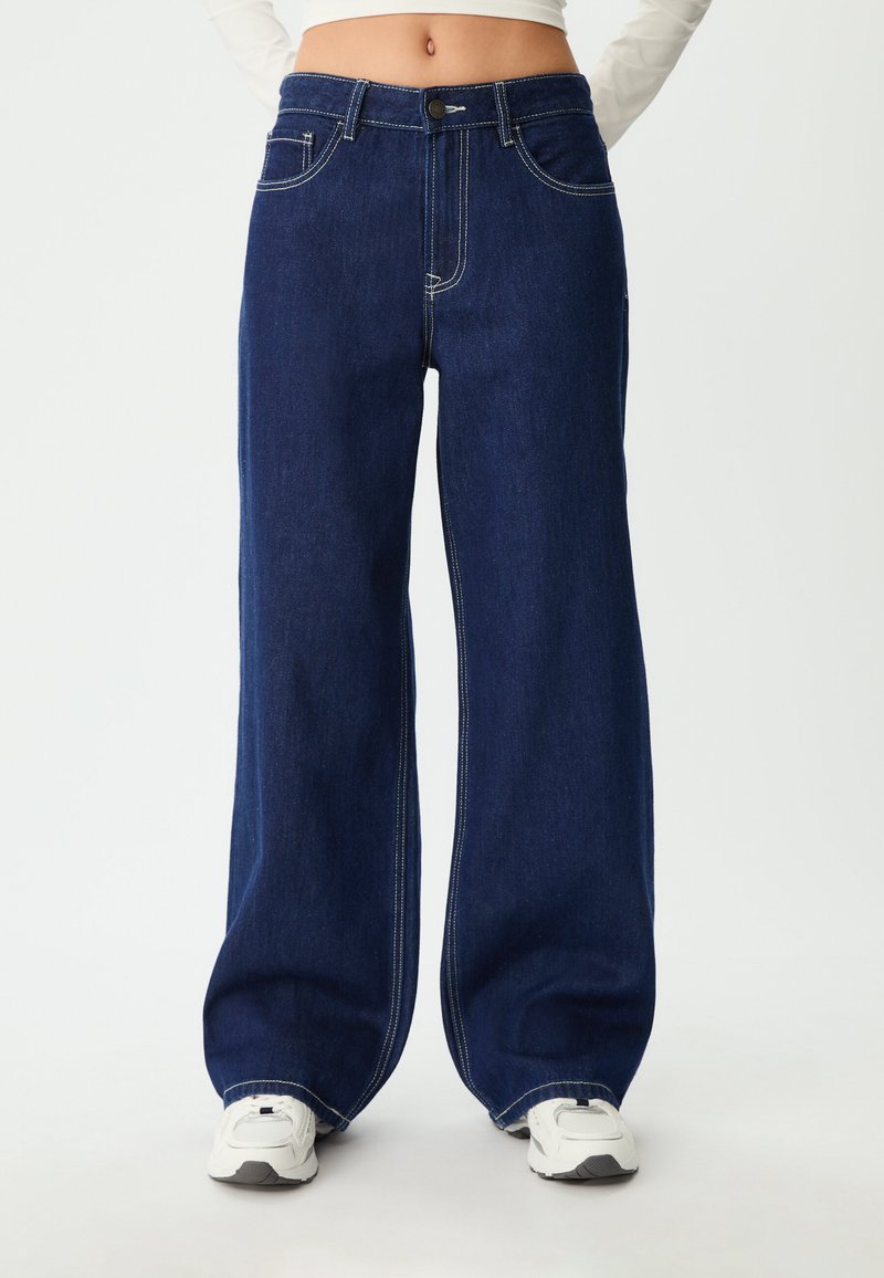 Tamno plave široke traperice od denima; imaju gustu šavovu obradu, dugme za zatvaranje i dizajn s pet džepova. Uparen s bijelim tenisicama.