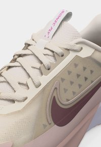 Κοντινό πλάνο του μπεζ και ροζ αθλητικού παπουτσιού Nike με ύφασμα από δίχτυ, πλευρική επιφάνεια με μοτίβο, ορατά κορδόνια και το κείμενο "SONIC FLY" στην γλώσσα.