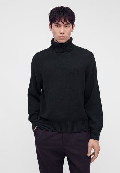 Jeune homme portant un pull à col roulé côtelé noir et un pantalon foncé, debout avec les mains partiellement dans les poches, sur un fond clair uni.