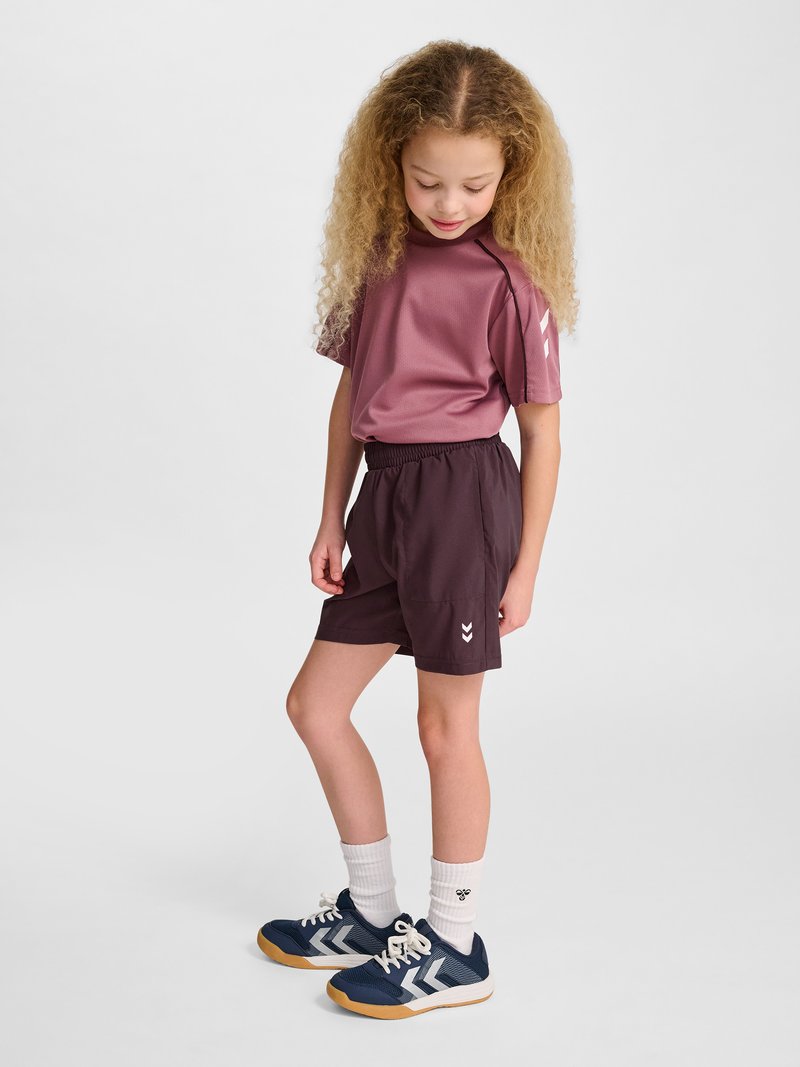 Ung pige med krøllet hår iført lilla sportstøj, marineblå sneakers og hvide sokker, står og kigger ned med et smil.