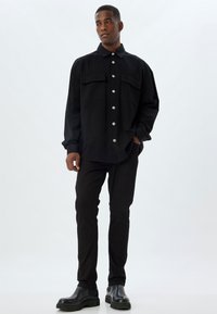 Musta paitajakku hopean värisillä napeilla, kaksi rintataskua, väljä malli, yhdistetty mustiin slim fit -housuihin ja tukeviin mustiin saappaisiin neutraalilla taustalla.