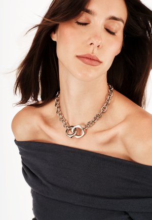 Sølvkæde med sammenlåste ringe, skinnende finish og teksturerede led. Model bærer en mørk off-shoulder top.