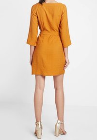 Kortärmad, orange klänning i texturerad tyg, med samlad midja och en avslappnad passform. Matchas med guldfärgade högklackade sandaler.