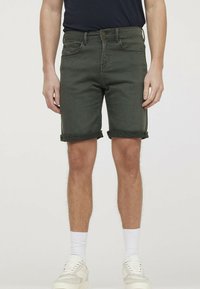 Shorts en denim vert olive pour homme avec revers repliés, design à cinq poches, coutures discrètes et coupe confortable. Baskets blanches et chaussettes montantes.