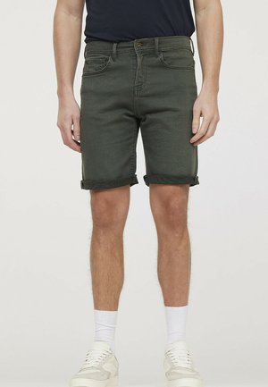 Herren Denim-Shorts in Olivgrün mit umgeschlagenen Säumen, Fünf-Taschen-Design, dezentem Faden und bequemer Passform. Weiße Sneaker und Crew-Socken.