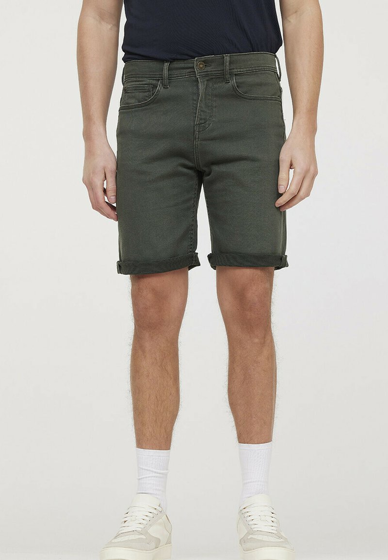 Shorts en denim vert olive pour homme avec revers repliés, design à cinq poches, coutures discrètes et coupe confortable. Baskets blanches et chaussettes montantes.
