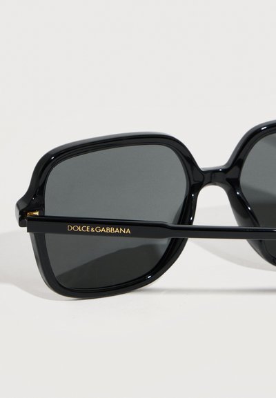 Μαύρα γυαλιά ηλίου με τετράγωνο σκελετό και φακούς με σκούρο χρώμα, με το λογότυπο "Dolce & Gabbana" σε χρυσό στην πλευρική βραχίονα, πάνω σε λευκό φόντο.