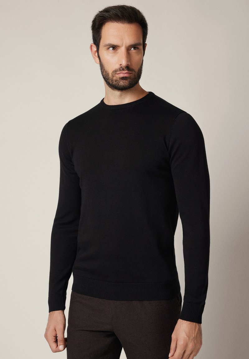 Mężczyzna o krótkich ciemnych włosach i brodzie, ubrany w dopasowany czarny sweter z długim rękawem i ciemne spodnie na jednolitym beżowym tle.