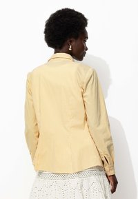 Chemise à rayures jaunes et blanches avec une coupe ajustée, manches longues et poignets fendus. Tissu lisse, col classique et détail de pli au dos.