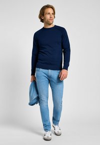 Granatowy sweter z dzianiny z prążkowanym dekoltem i mankietami, w połączeniu z jasnoniebieskimi dżinsami i białymi sneakersami. Na ramieniu zawieszona kurtka dżinsowa.
