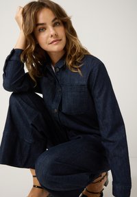 Junge Frau mit welligem Haar, die eine dunkelblaue Jeansjacke und -hose trägt, in hockender Position mit der Hand an ihrem Kopf vor einem einfachen Hintergrund.