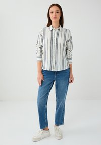 Hemd mit Knöpfen, vertikalen blauen und grauen Streifen auf weißem Hintergrund. Kombiniert mit blauen Jeans und weißen Sneakers.
