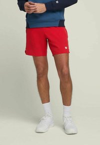Persona vestida con pantalones cortos deportivos rojos, calcetines blancos y zapatillas blancas, de pie con las manos entrelazadas, frente a un fondo claro y liso.