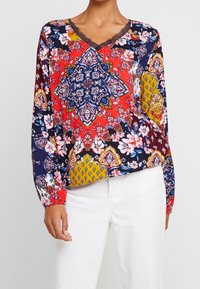 Pitkähihaista puseroa, jossa on V-kaula-aukko. Monivärinen kukkakuvio ja geometriset kuviot punaisella, laivastonsinisellä ja keltaisella taustalla. Hieno pitsireunus.