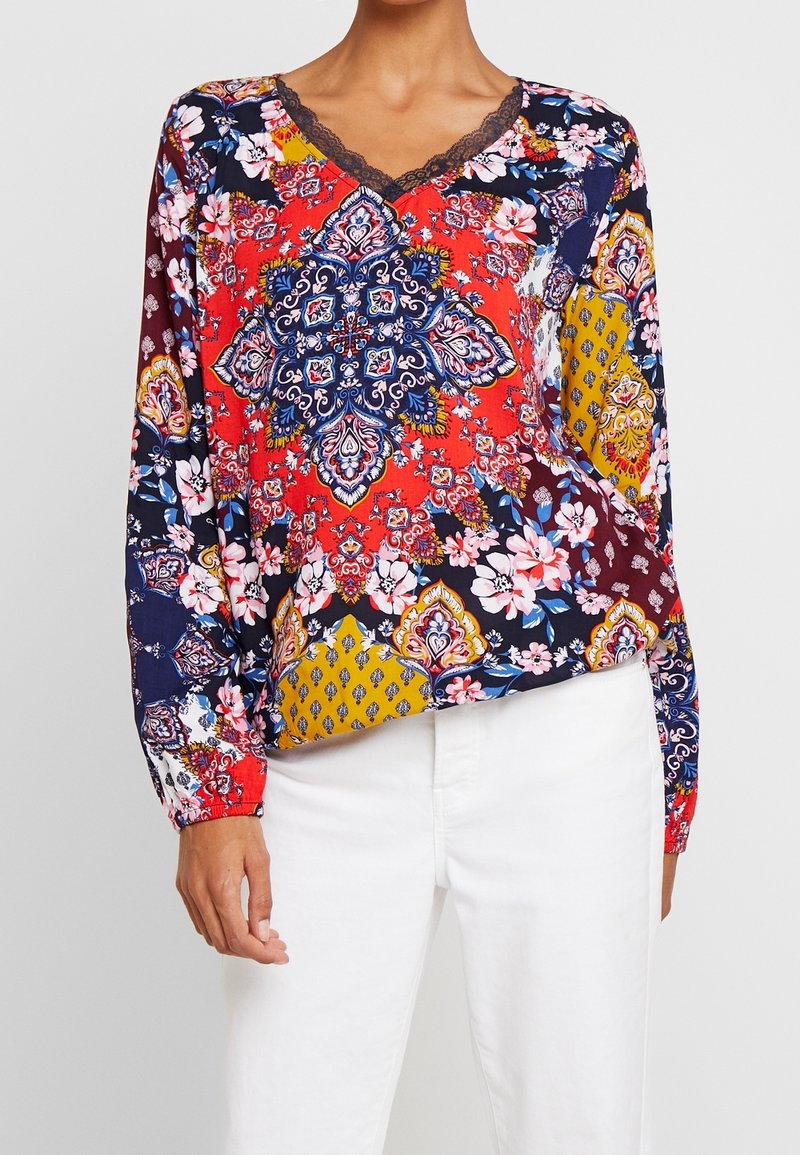 Pitkähihaista puseroa, jossa on V-kaula-aukko. Monivärinen kukkakuvio ja geometriset kuviot punaisella, laivastonsinisellä ja keltaisella taustalla. Hieno pitsireunus.