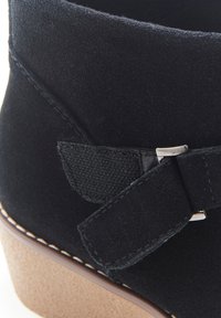 Botín de ante negro con un lazo texturizado y herrajes plateados, que presenta una suela de plataforma marrón clara y costuras blancas en contraste.