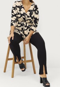 Zwarte en beige bloementunika met een V-hals en een cinched taille, gecombineerd met zwarte leggings met inkepingen bij de enkels en zwarte blokhak sandalen.