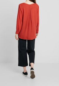 Blouse rouge à manches longues avec un col arrondi et un pli au dos, associée à un pantalon noir à jambes larges et des chaussures noires. Texture de tissu lisse.
