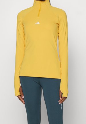 Person iført en gul langærmet træningstrøje med hul til tommelfingrene og tætsiddende turkise leggings med mærket "Techfit".