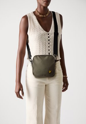 Olive green square bag with a textured finish, black strap, and yellow logo detail. Worn crossbody against a light background.
