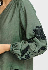 Blusa verde con un tessuto testurizzato. Presenta maniche a sbuffo e motivi di foglie ricamati in nero sulla manica sinistra.