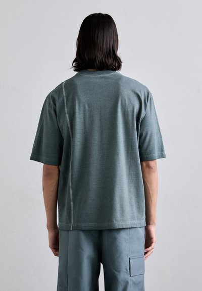 T-shirt ample à manches courtes en tissu vert doux. Présente une surface texturée et des coutures contrastantes le long de la couture arrière.