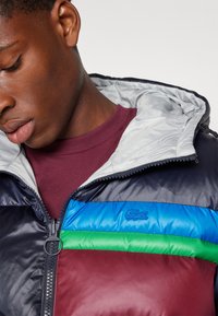 Chaqueta acolchada en navy y burdeos con un forro gris. Presenta rayas horizontales en azul y verde, un cierre con cremallera y detalles de logotipo bordado.