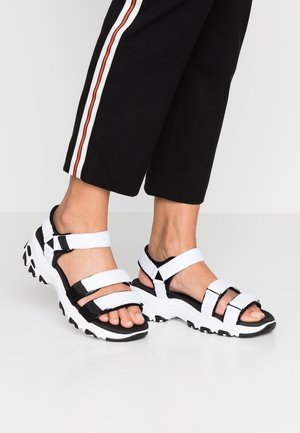 Fødder iført sorte og hvide sportslige sandaler med tykke såler, kombineret med sorte bukser med hvide og røde sidelommer.