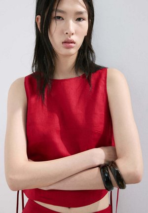 Young woman with long dark hair wearing a sleeveless red top, arms crossed, and two dark bangles on her right wrist against a plain background.