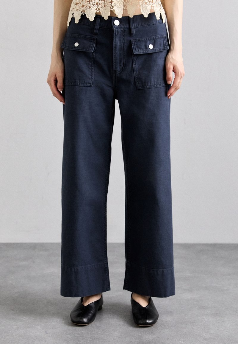 Jean large marine en denim de coton avec poches latérales et ourlet brut. Porté avec un haut en dentelle et des chaussures noires sans lacets.