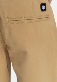 Tan katoenen broek met een achterzak die een zwart logo-patch en stiksels heeft. Glad textuur met een getailleerde pasvorm.