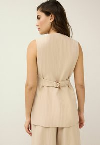 Gilet sans manches beige avec des rayures verticales, ceinturé à la taille avec une boucle circulaire, présentant une texture de tissu lisse.